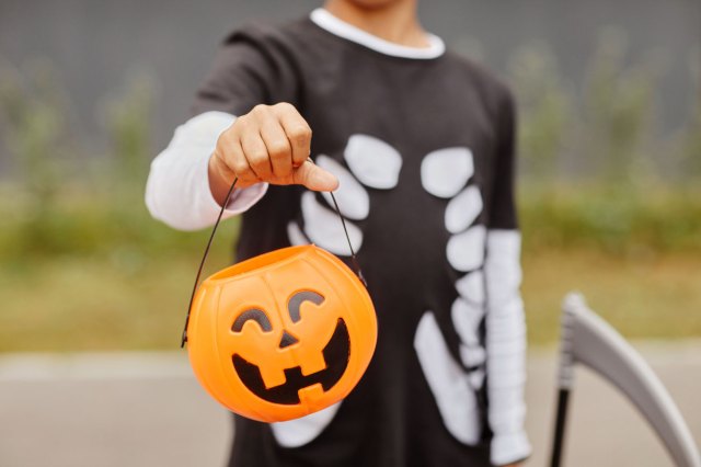 An image of a kid in a skeleton costume holding a jack-o'-lantern candy bucket