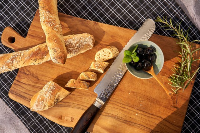 An image of loaves and slices of bread on a wooden cutting board with a knife and a bowl of olives