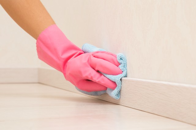 An image of a hand wearing a pink rubber glove cleaning a baseboard with a light blue cloth